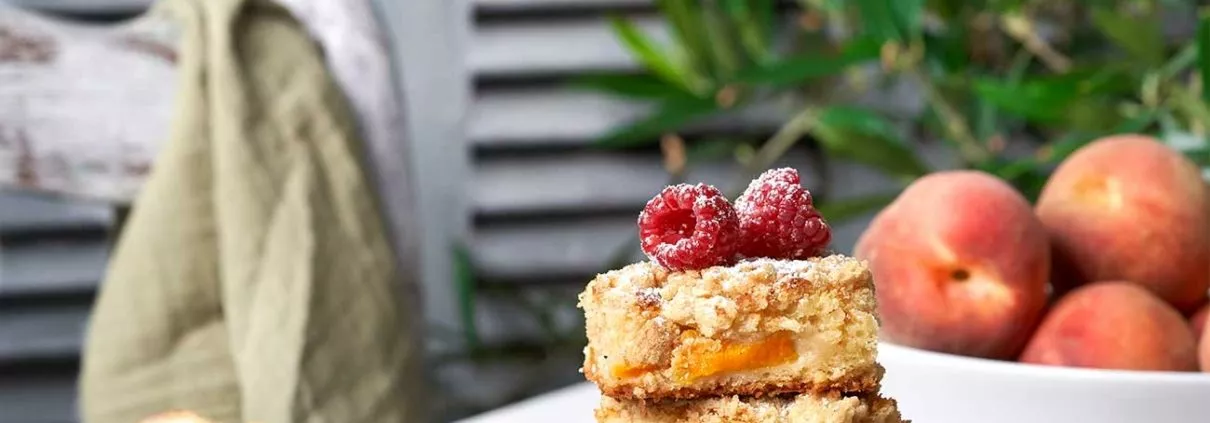 Pfirsichkuchen angerichtet auf einem weißen Tisch mit Pfirsichen im Hintergrund