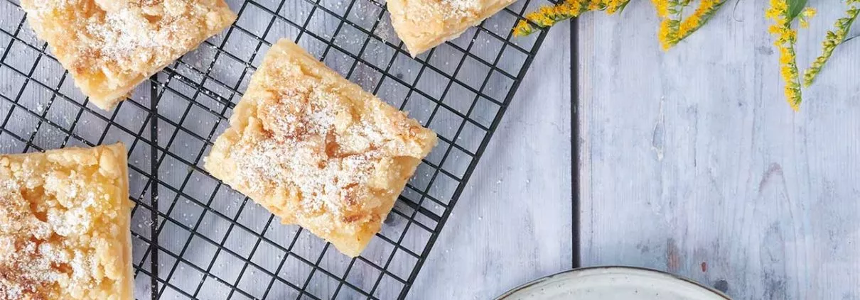 Apfelmus Streuselkuchen mit Puderzucker bestäubt auf einem blauen Holztisch mit gelben Blumen